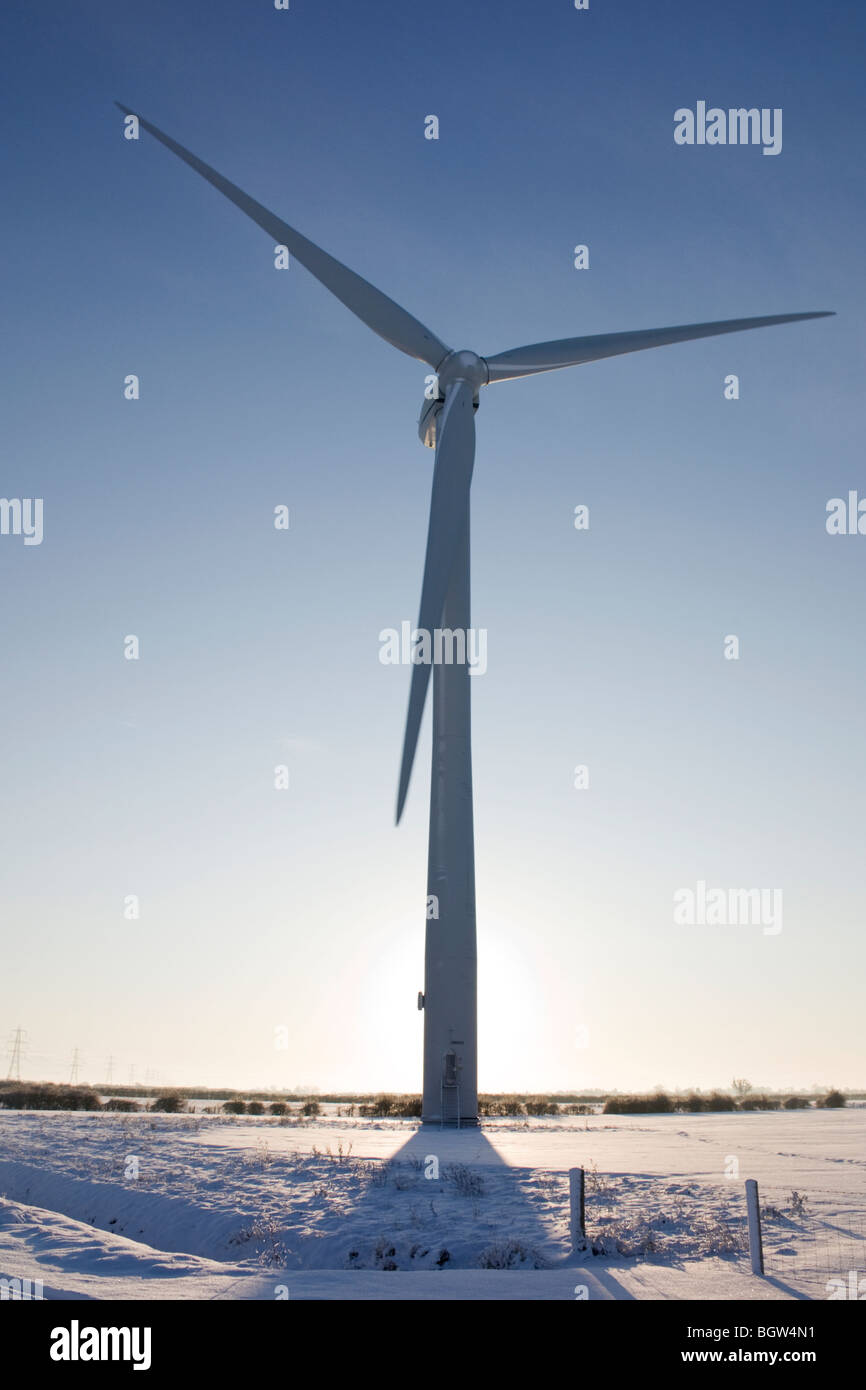 Wind turbine in snow dominating a flat fenland landscape and silhouetted against the sunrise in a blue winter sky Stock Photo