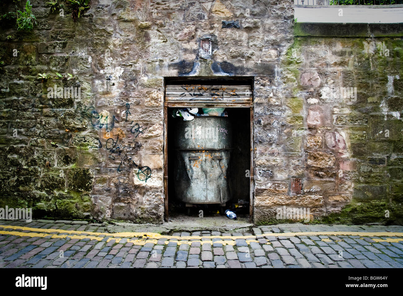 A metal bin in a recess within an old brick wall with a rusty metal shutter Stock Photo