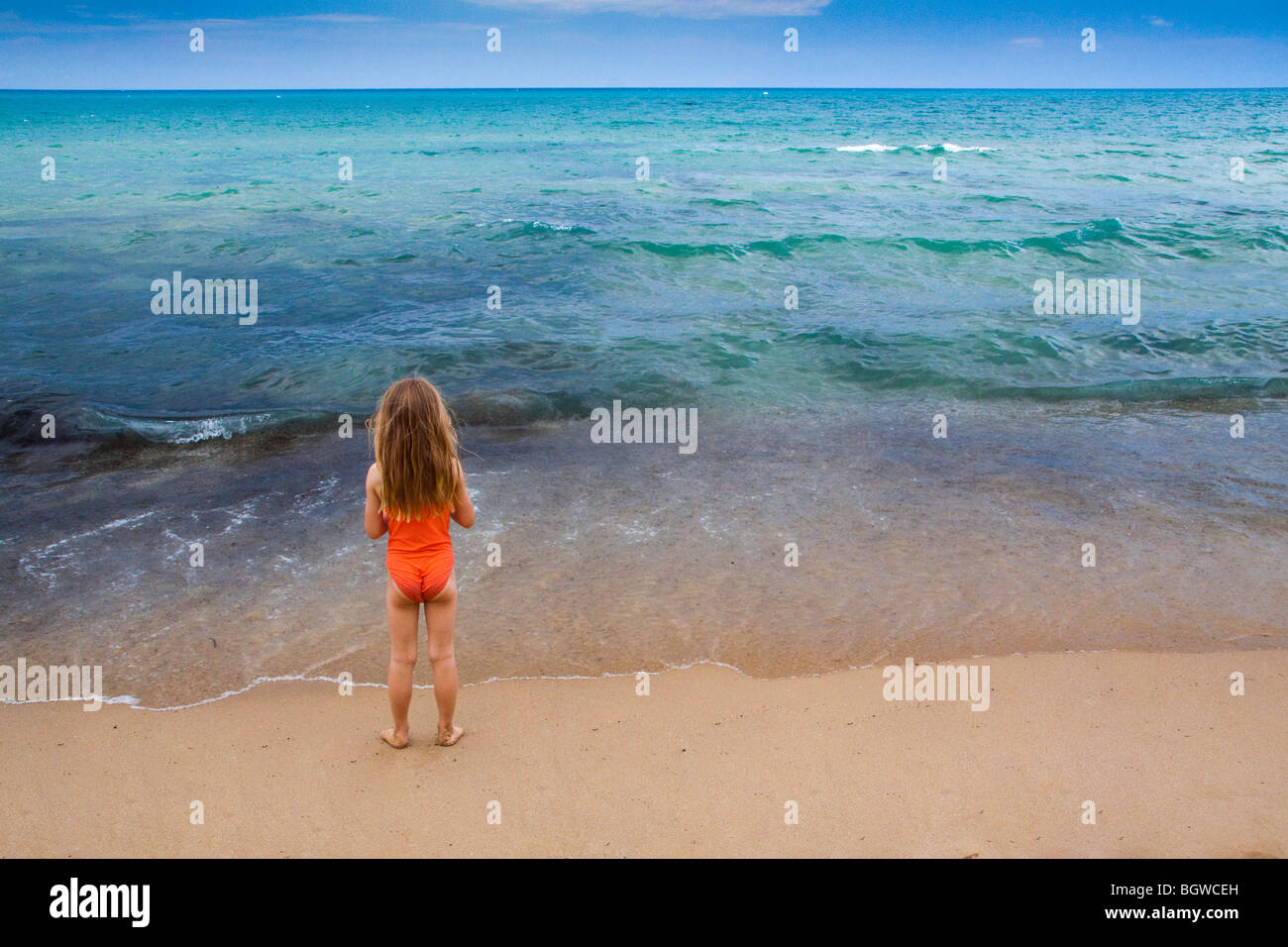 Young girl in swimsuit with long hair waits on beach, looking out to sea Stock Photo