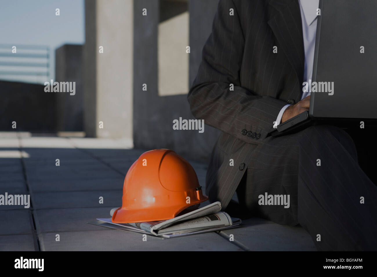 Man sitting on sidewalk using laptop, orange hard hat set on newspaper