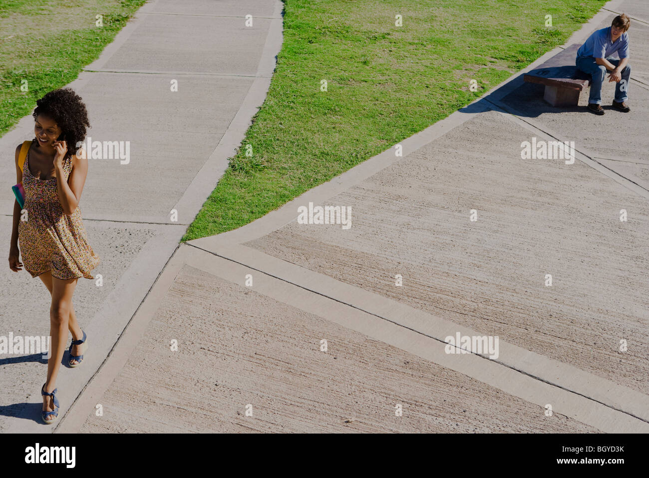 Attractive young woman walking in park, talking on cell phone, young man watching her from nearby bench Stock Photo
