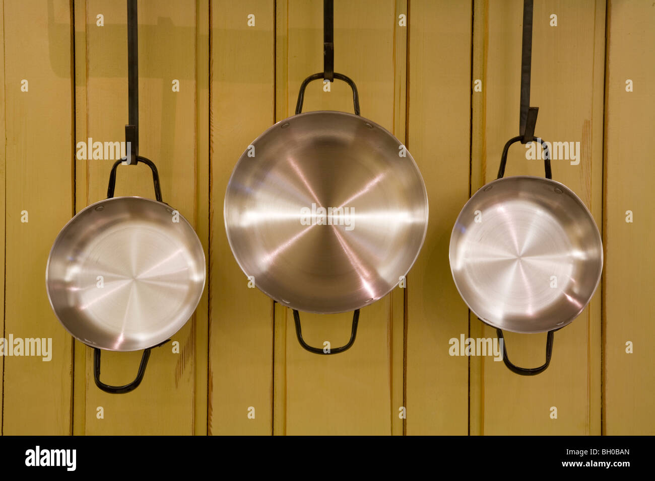 Three frying pans casseroles griddles poachers hanging from metal grips in front of yellow wooden wall in kitchen Stock Photo