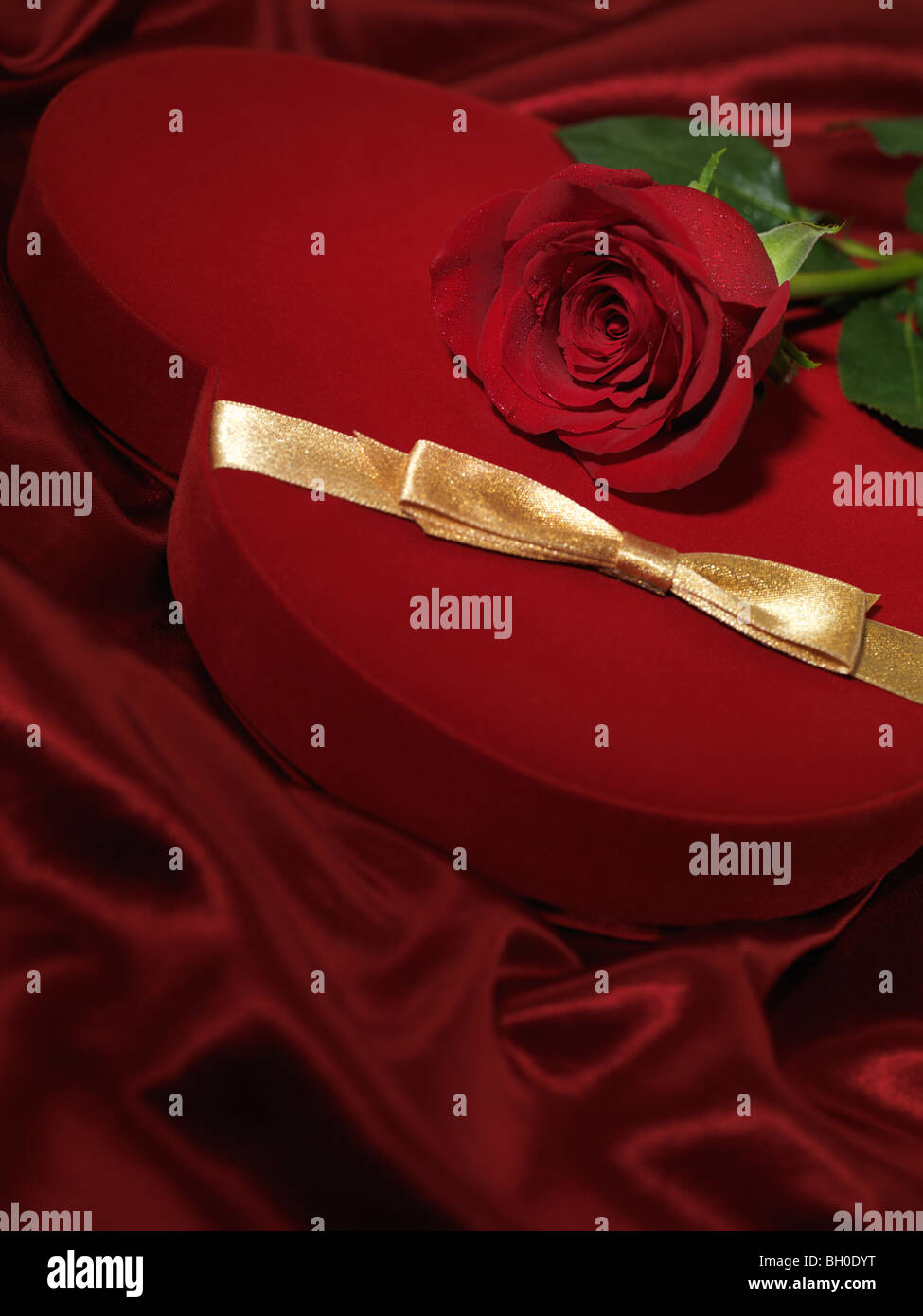 Red heart-shaped gift box and a red rose on red silky cloth Stock Photo