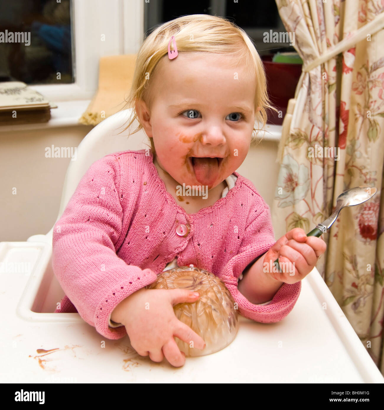Square close up portrait of a baby girl getting in a mess eating chocolate ice cream in her high chair. Stock Photo