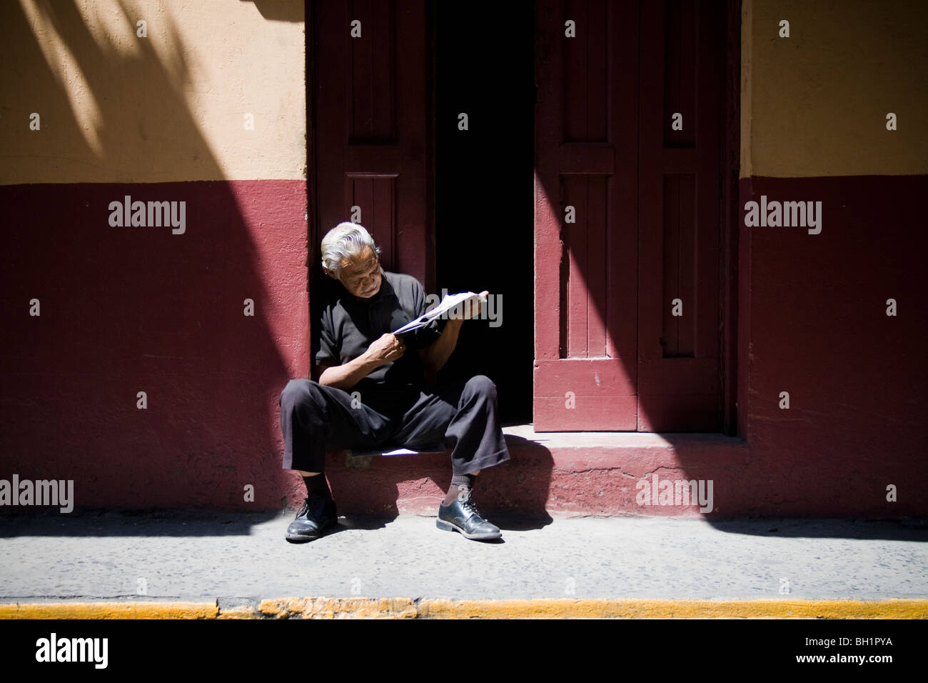 An old man sits to read the newspaper on his doorstep on a quiet street in the old town area of Panama city Stock Photo