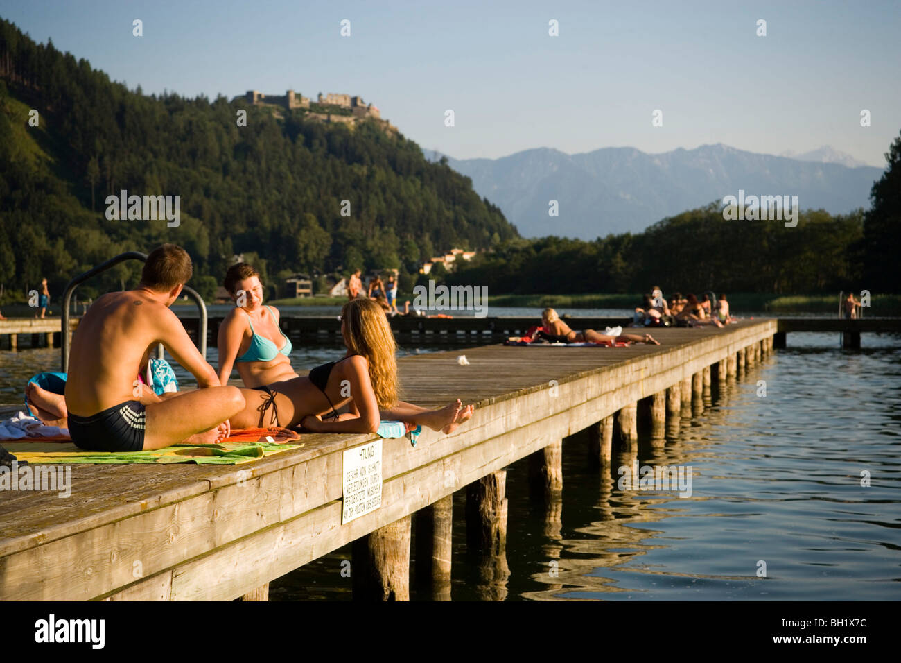 Young people sunbathing on boardwalk, Castle Landskron in background, Lake Ossiach, Ossiach, Carinthia, Austria Stock Photo