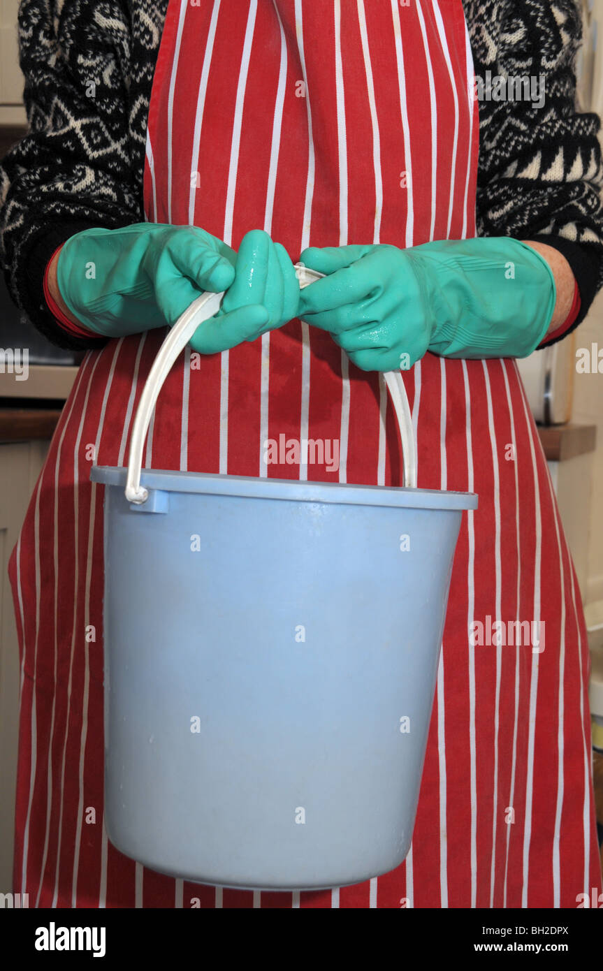 A woman wearing an apron and with rubber gloves on her hands carries a blue pail. Stock Photo