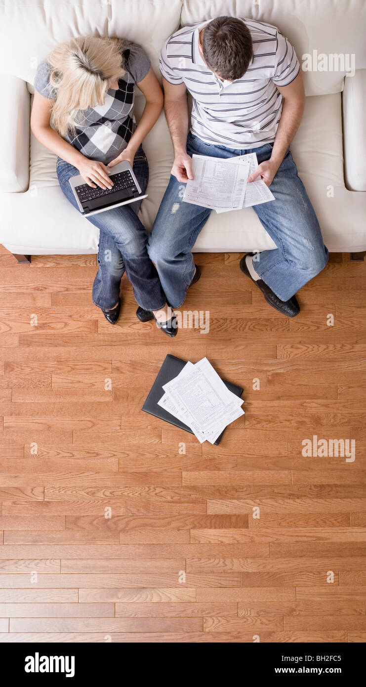 High angle view of young couple sitting on a couch with a laptop and paperwork. Vertical shot. Stock Photo