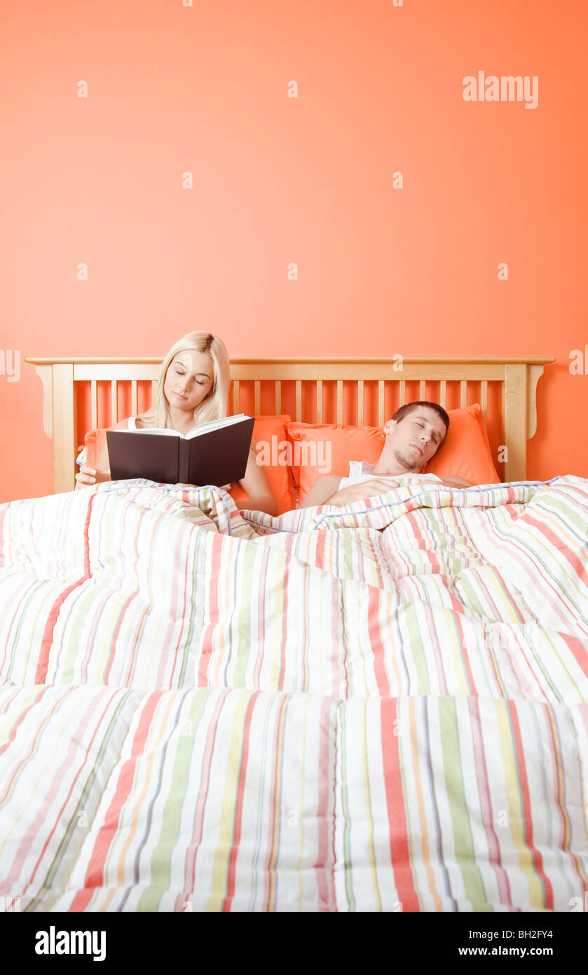 View of couple in bed, with woman reading book and man sleeping
