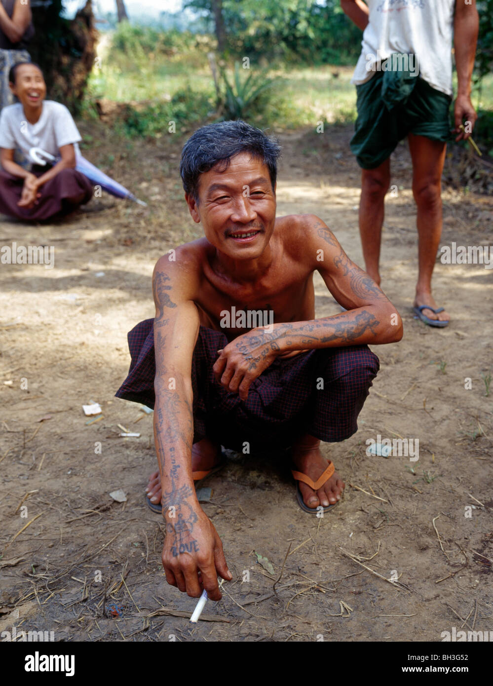 squatting tattooed Burmese smoking a cigarette during a rest tätowierter Burmese raucht Zigarette in einer Pause Myanmar Burma Stock Photo