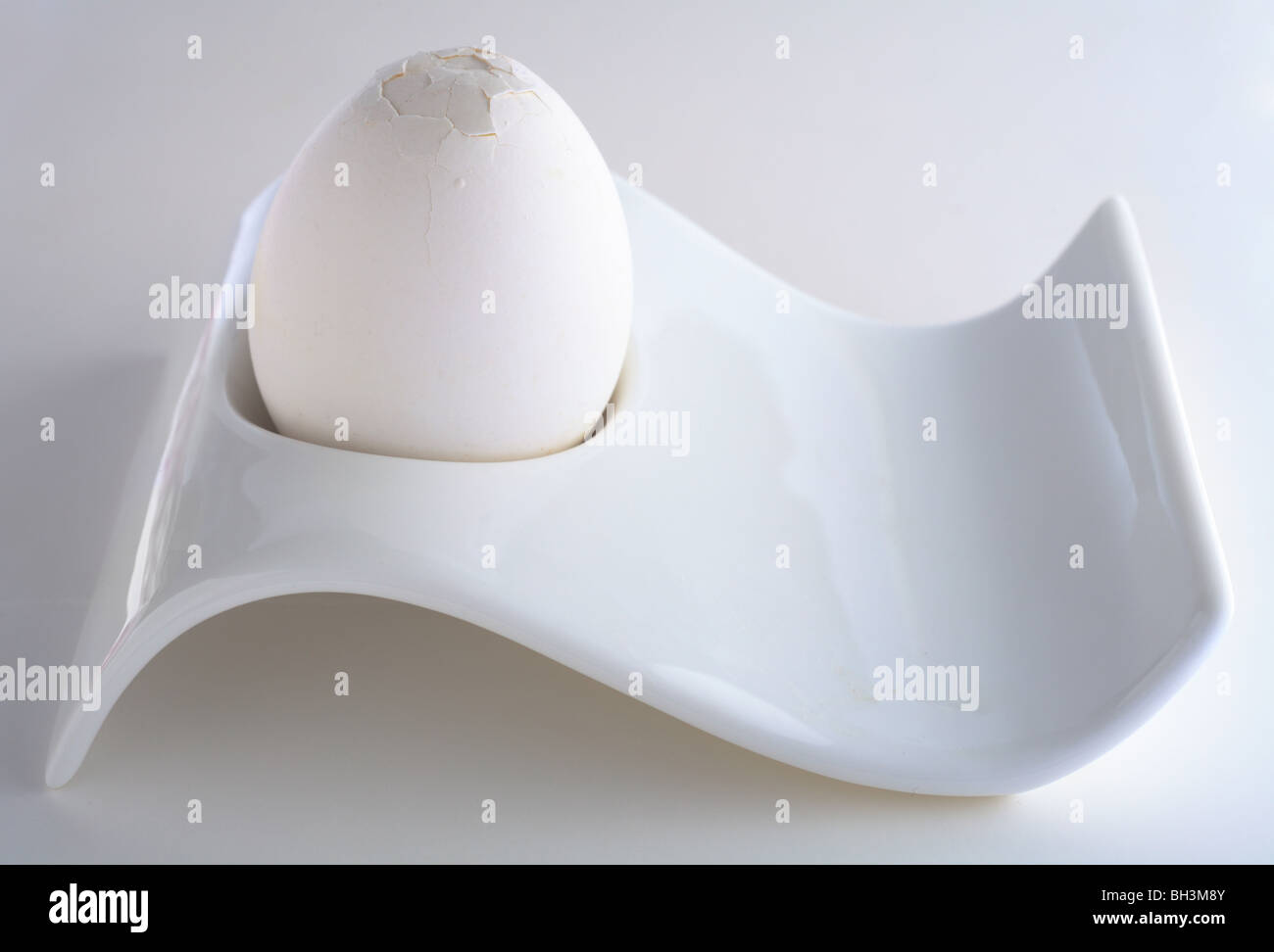 A white boiled egg with its top cracked ready for peeling in a white eggcup on a plain background, with a soft shadow Stock Photo
