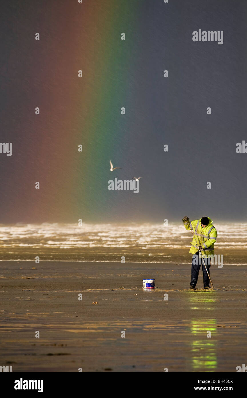 Cockle picker in yellow jacket and rainbow in background over sea Stock Photo