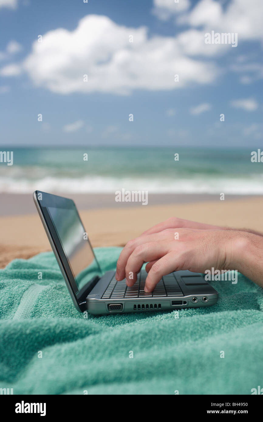 A man's hands working on a laptop computer on a beach towel on a deserted beach Stock Photo