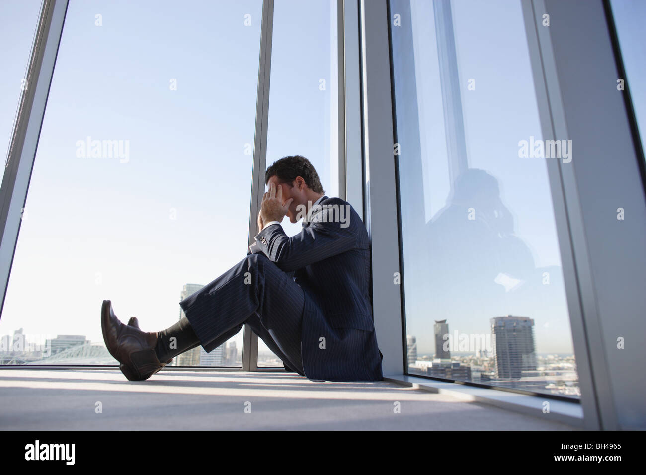 A businessman sitting on the floor in the corner of an office skyscraper with his head in his hands Stock Photo