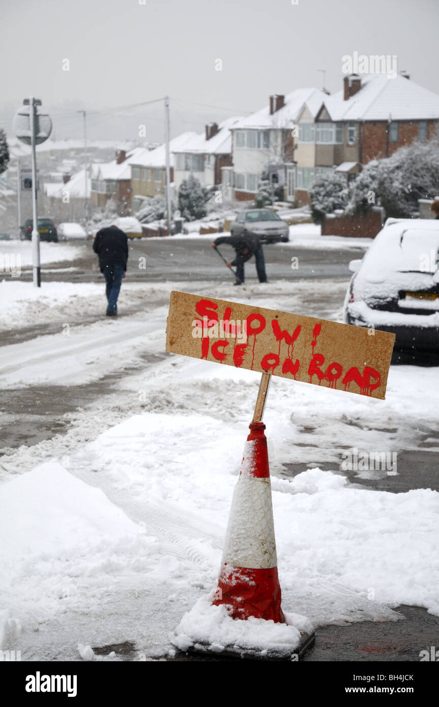 Makeshift signpost warning of ice on on suburban road and men in the background clearing compacted snow. Stock Photo