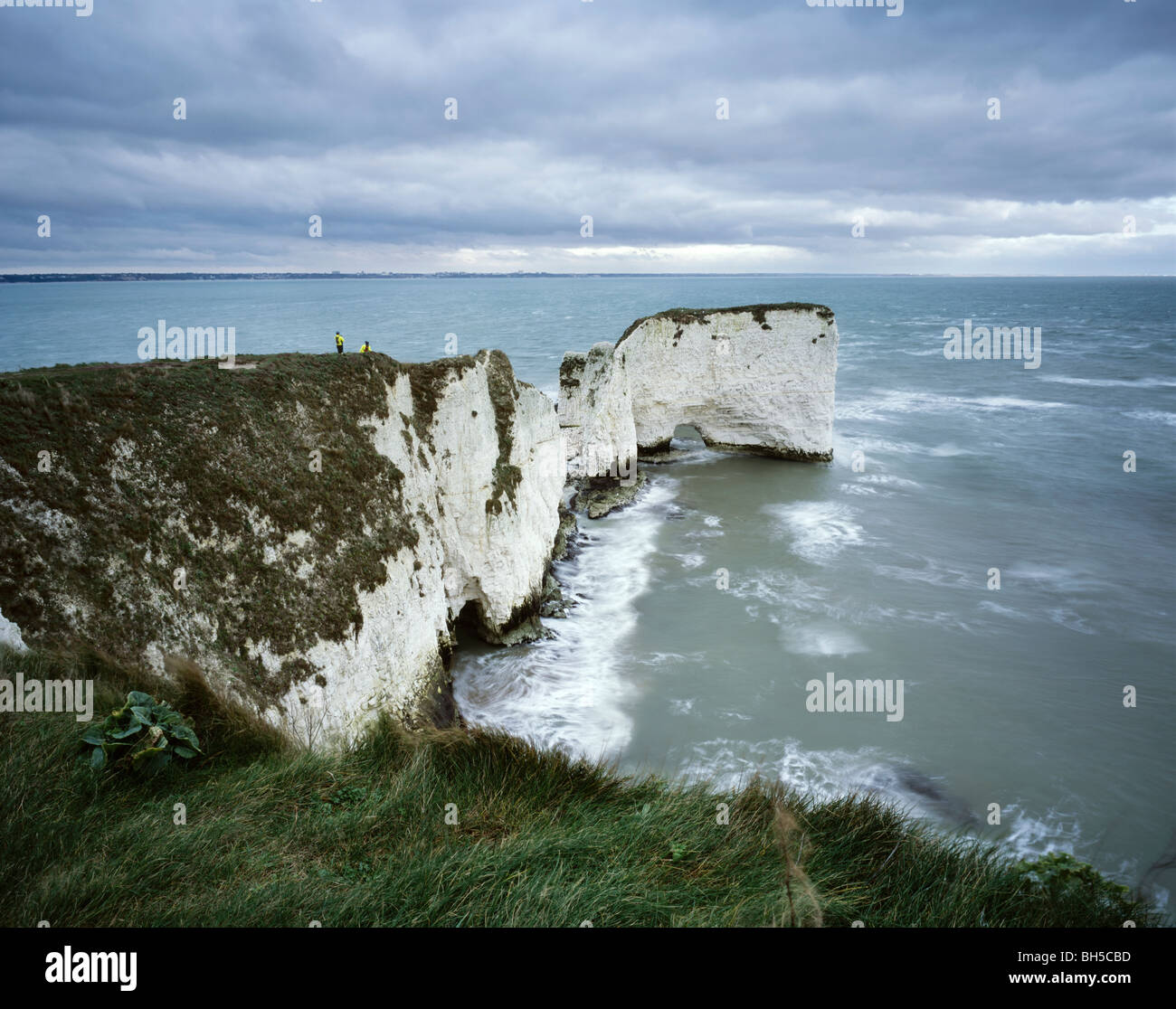Classic view of Old Harry Rocks in Dorset on a cloudy day with 2 people near the edge of the rock giving an idea of the scale. Stock Photo