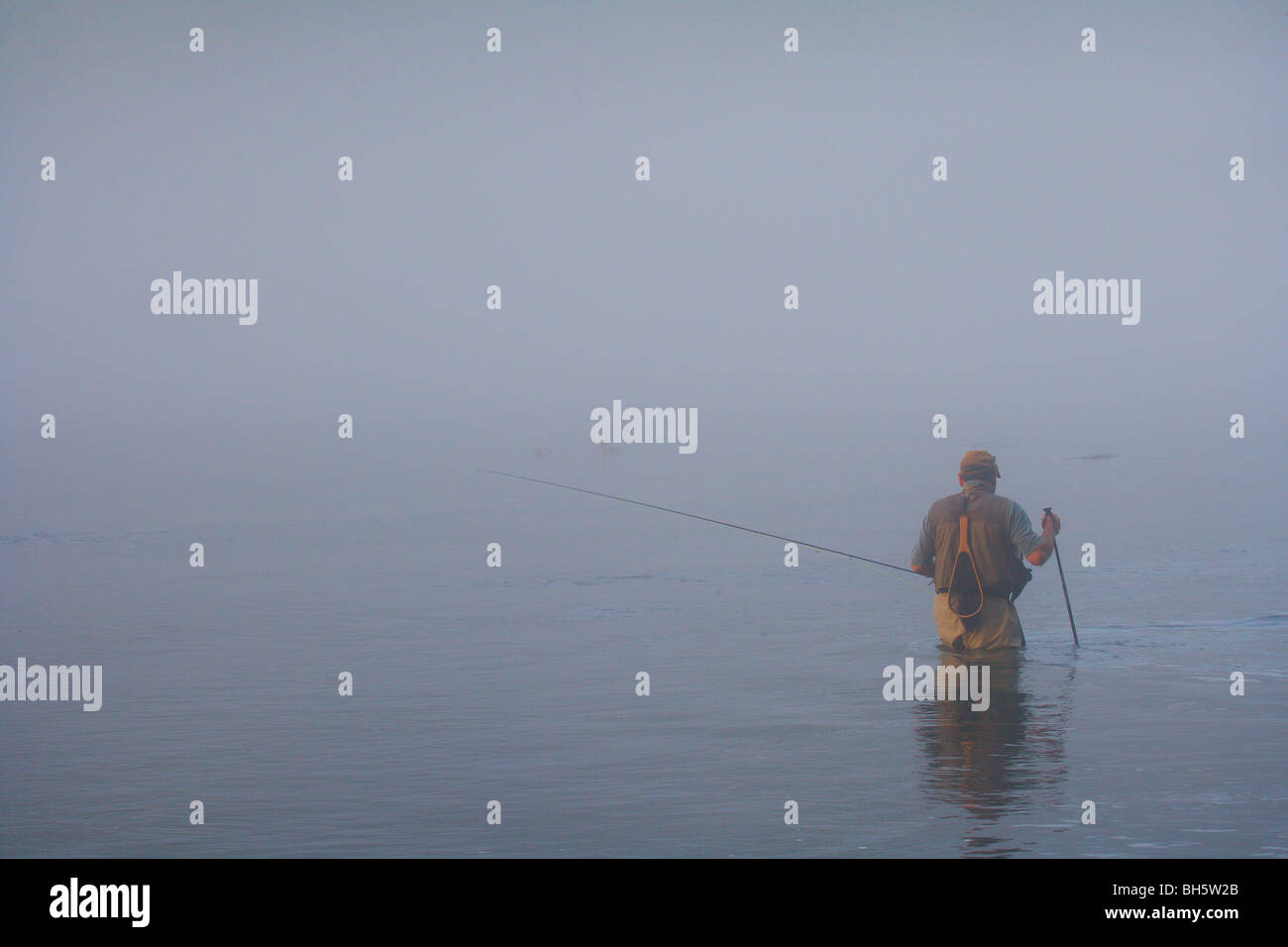 FLY FISHERMAN WADING IN RIVER CARRYING FLY ROD HEAVY FOG BAD WEATHER Stock Photo