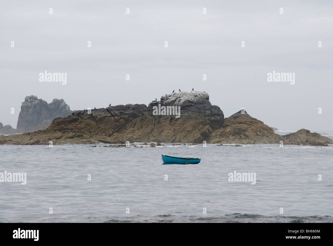 Single blue fishing boat floating on the sea against background of rocks and grey sky - Cornwall, UK Stock Photo