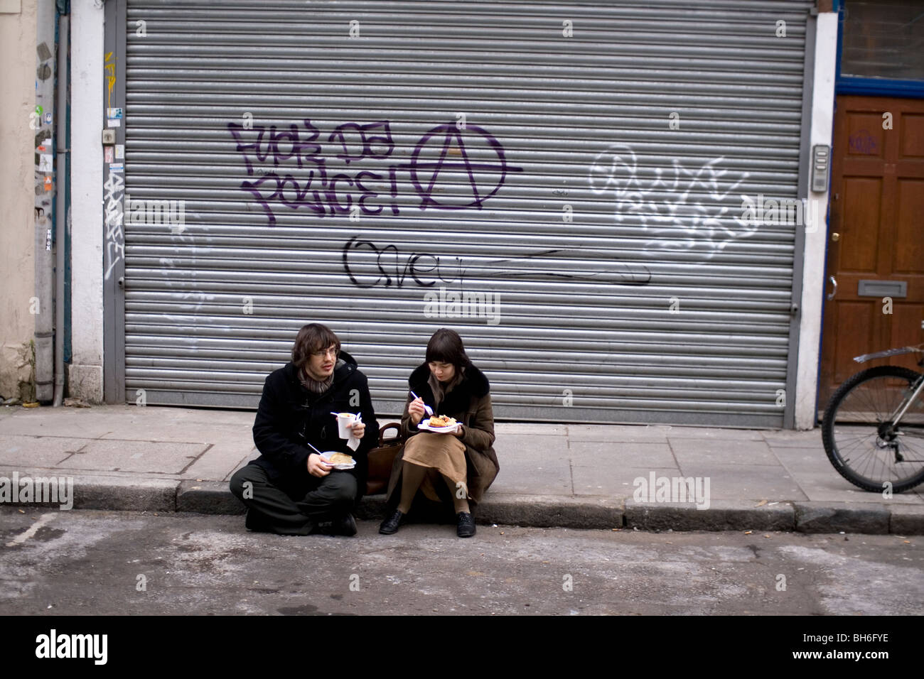 A couple sit and eat their take-away while sitting on a curb in London. Stock Photo