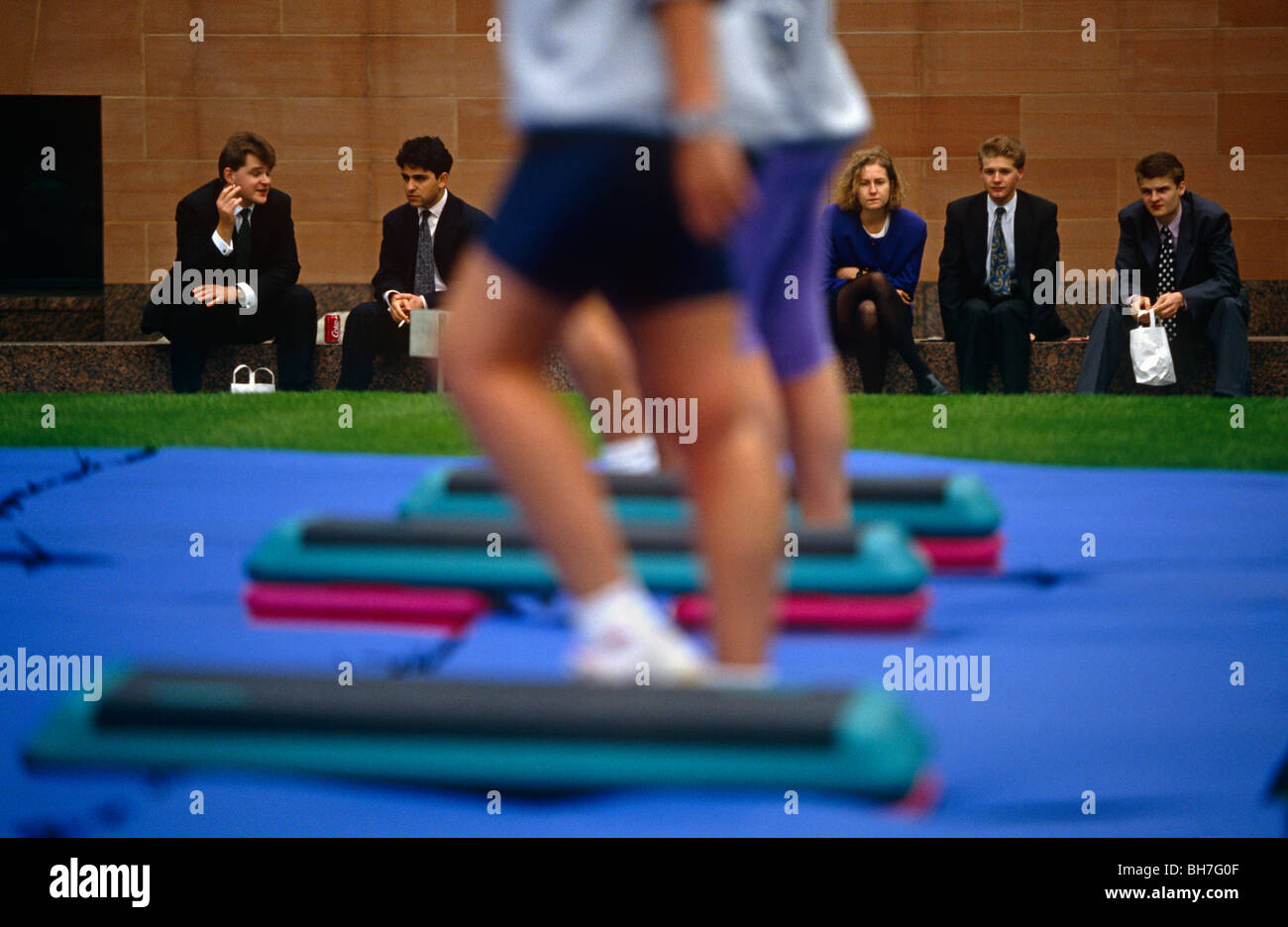 Guilty-looking office workers watch a group of fitness fanatics demonstrate exercises during a lunchtime promotion in Broadgate Stock Photo