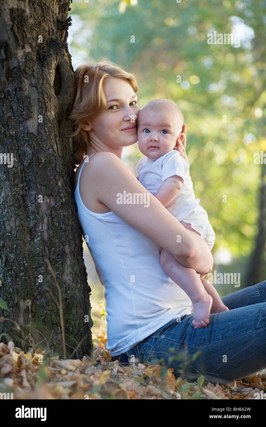 A mother sitting against a tree kissing her baby, looking at camera Stock Photo