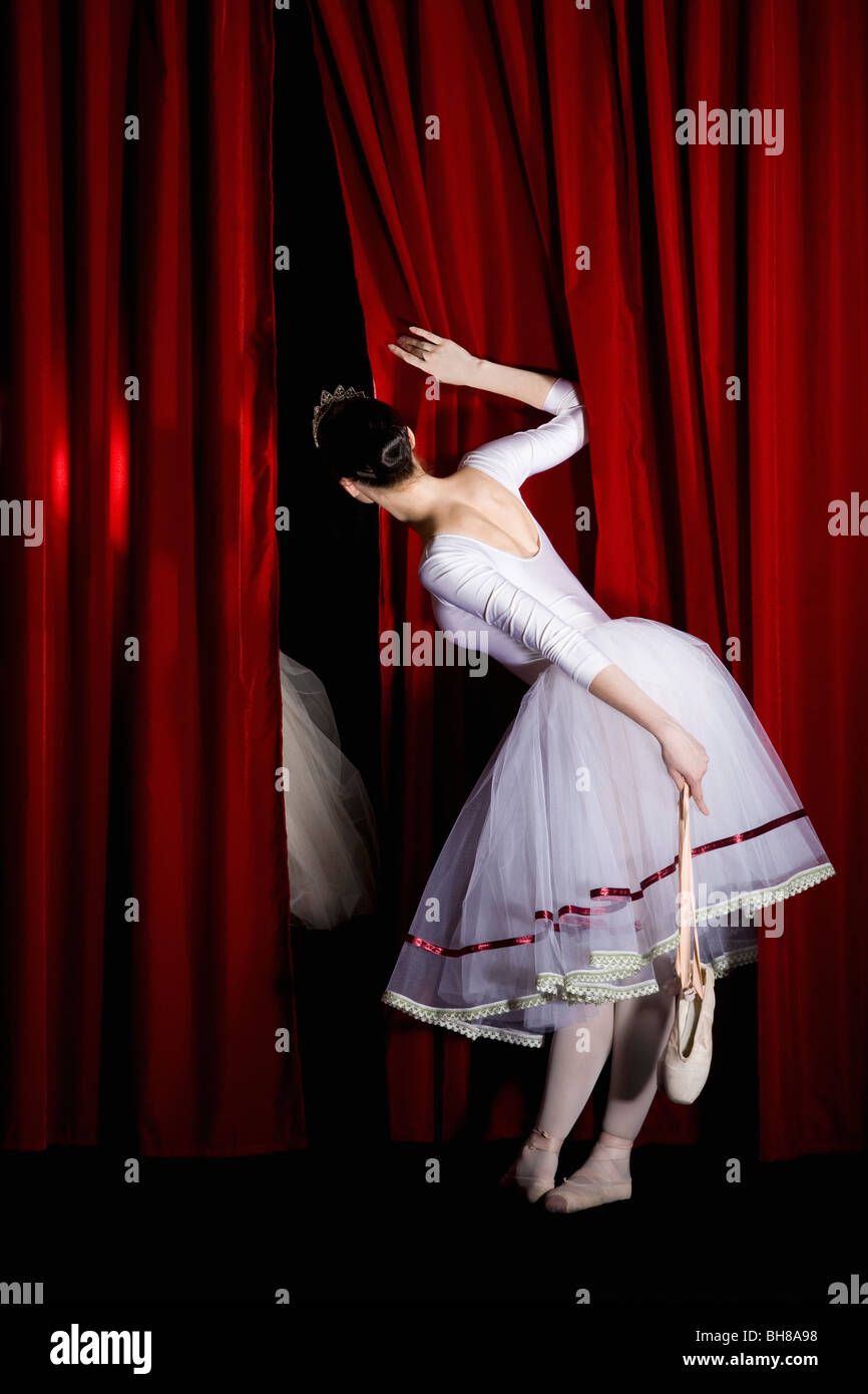 A ballet dancer peeking through a stage curtain holding a pointe shoe, rear view Stock Photo