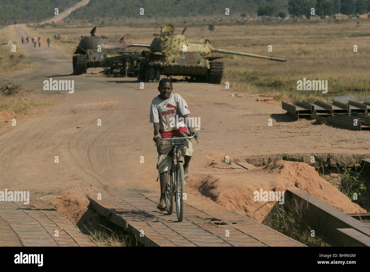 Angolan boy on bicycle rides past two Cuban tanks which were abandoned at Longa  in Southern Angola, Africa. Stock Photo