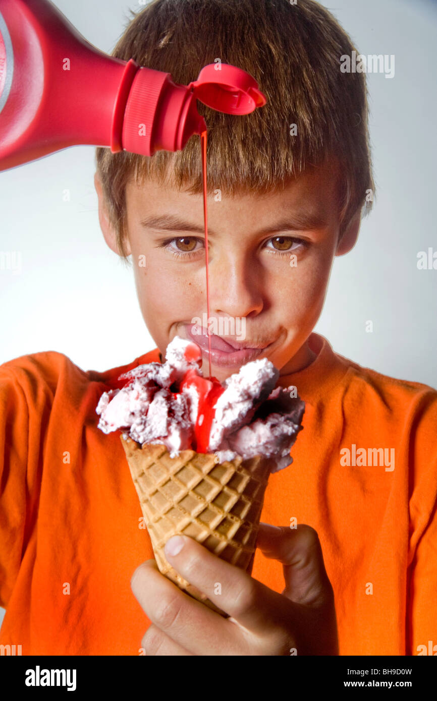 A happy ten year old boy pours strawberry syrup onto a scoop of ice cream in a waffle cone. Stock Photo