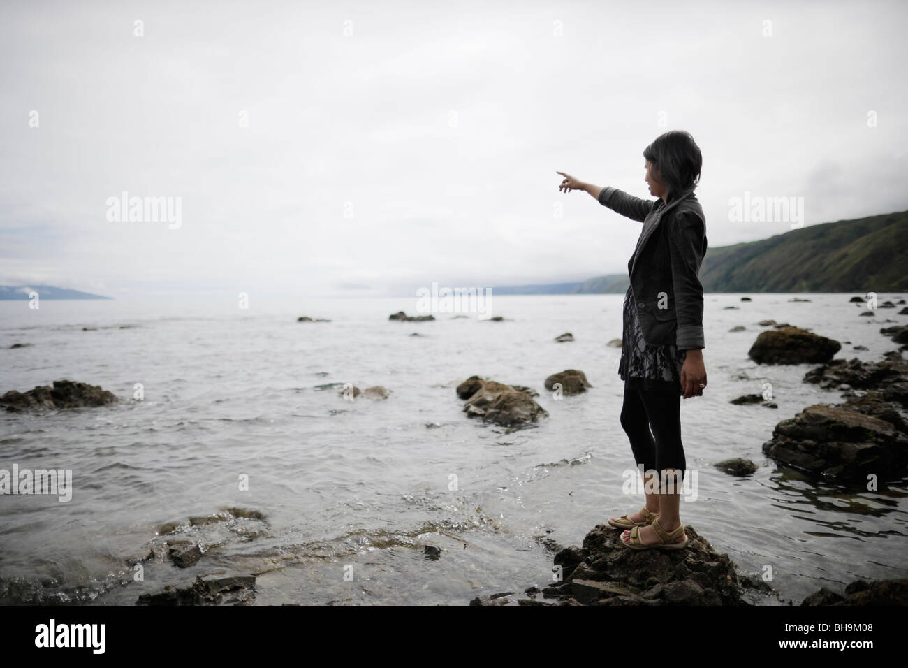 woman on rocky shore pointing out to sea Stock Photo