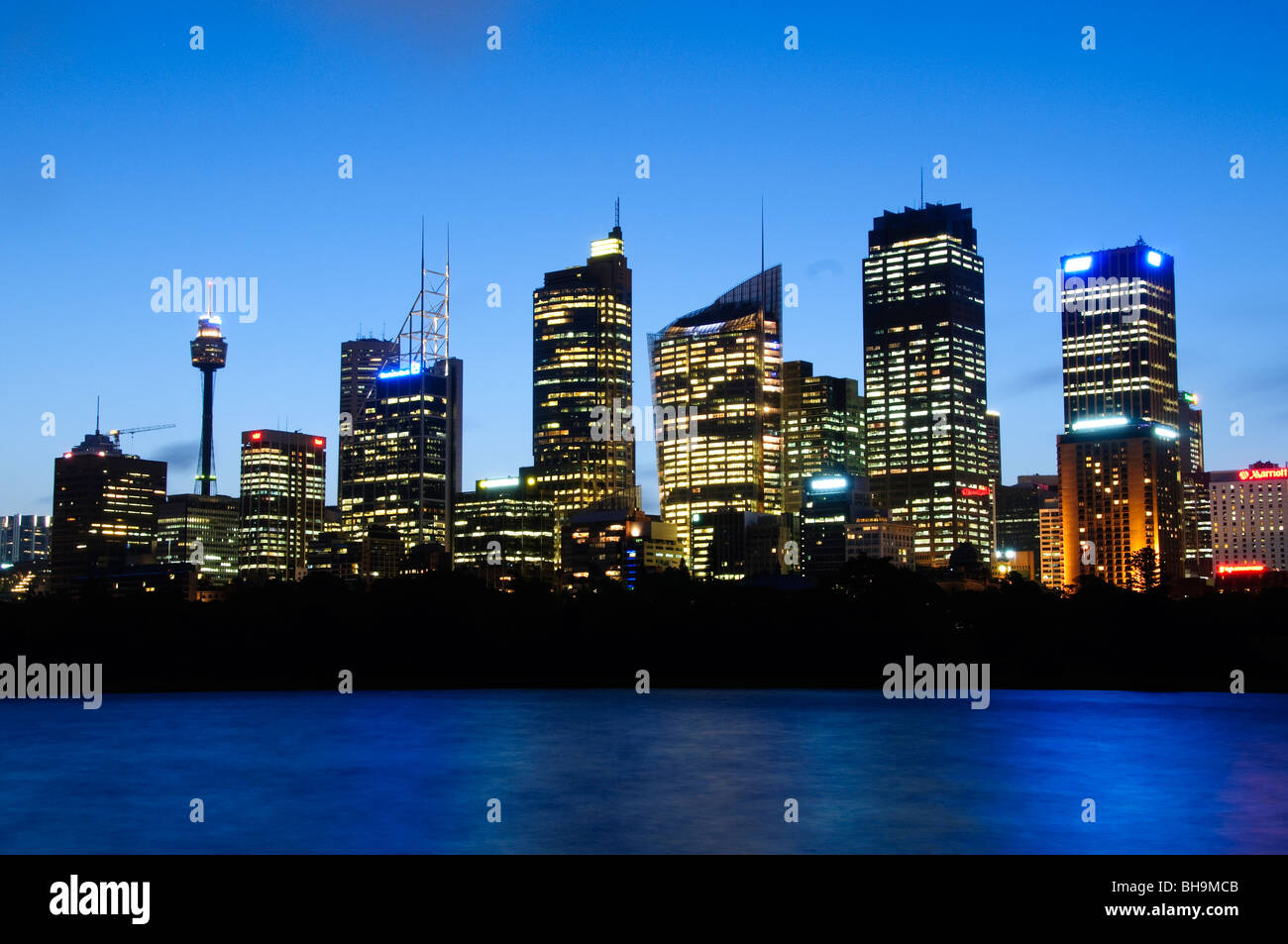 Sydney Skyline From Mrs Macquarie's Point Sydney Australia // SYDNEY, Australia — Dusk view of Sydney's city skyline as seen from Mrs. Macquarie's Point. The city's iconic buildings, including Sydney Tower, are brightly illuminated against the deep blue twilight sky, reflecting softly on the water in the foreground. This vantage point offers a classic perspective of the vibrant urban landscape. Stock Photo