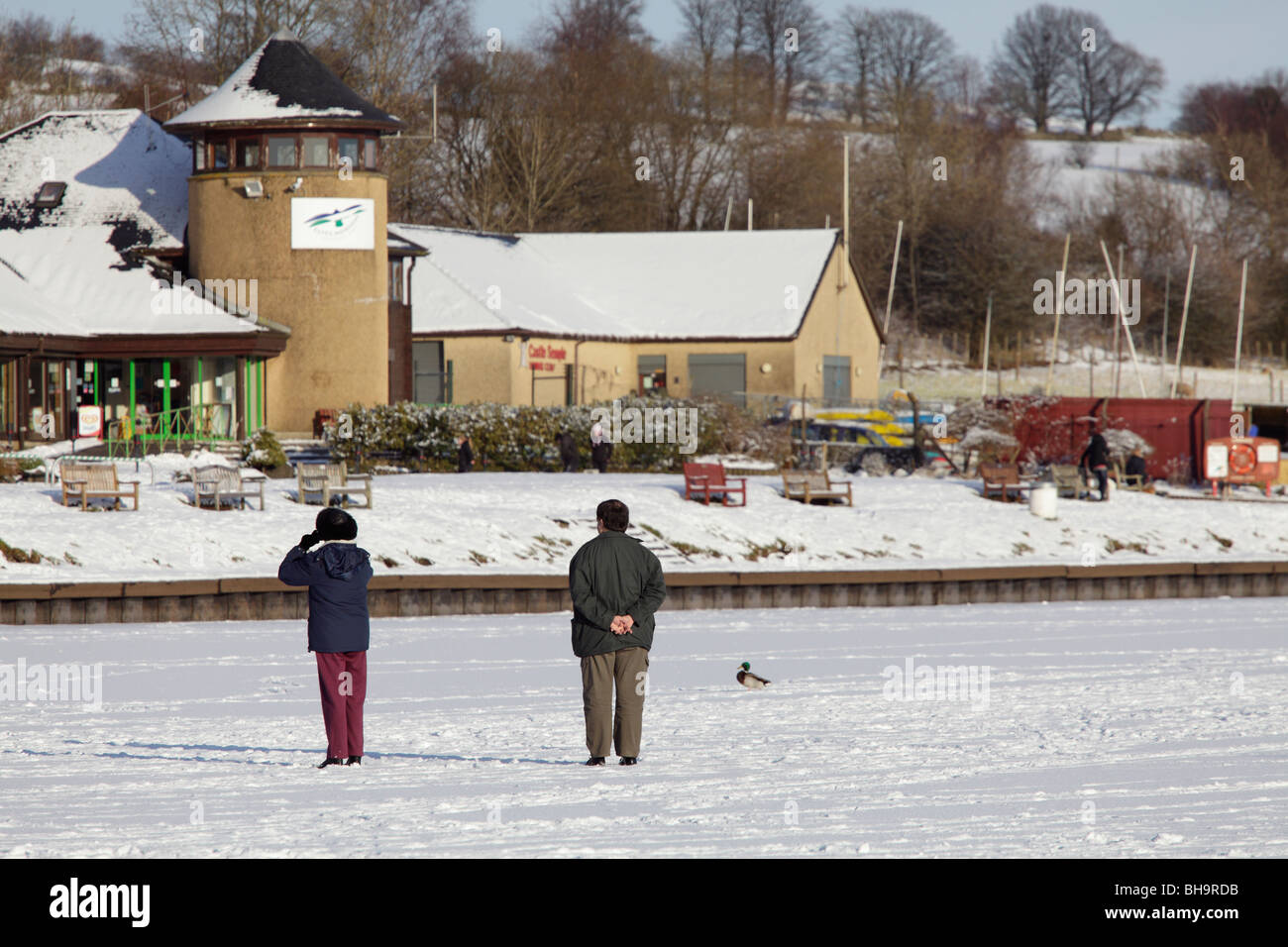 Couple standing on a frozen and snow covered Castle Semple Loch, Clyde Muirshiel Regional Park, Lochwinnoch, Renfrewshire, Scotland, UK Stock Photo