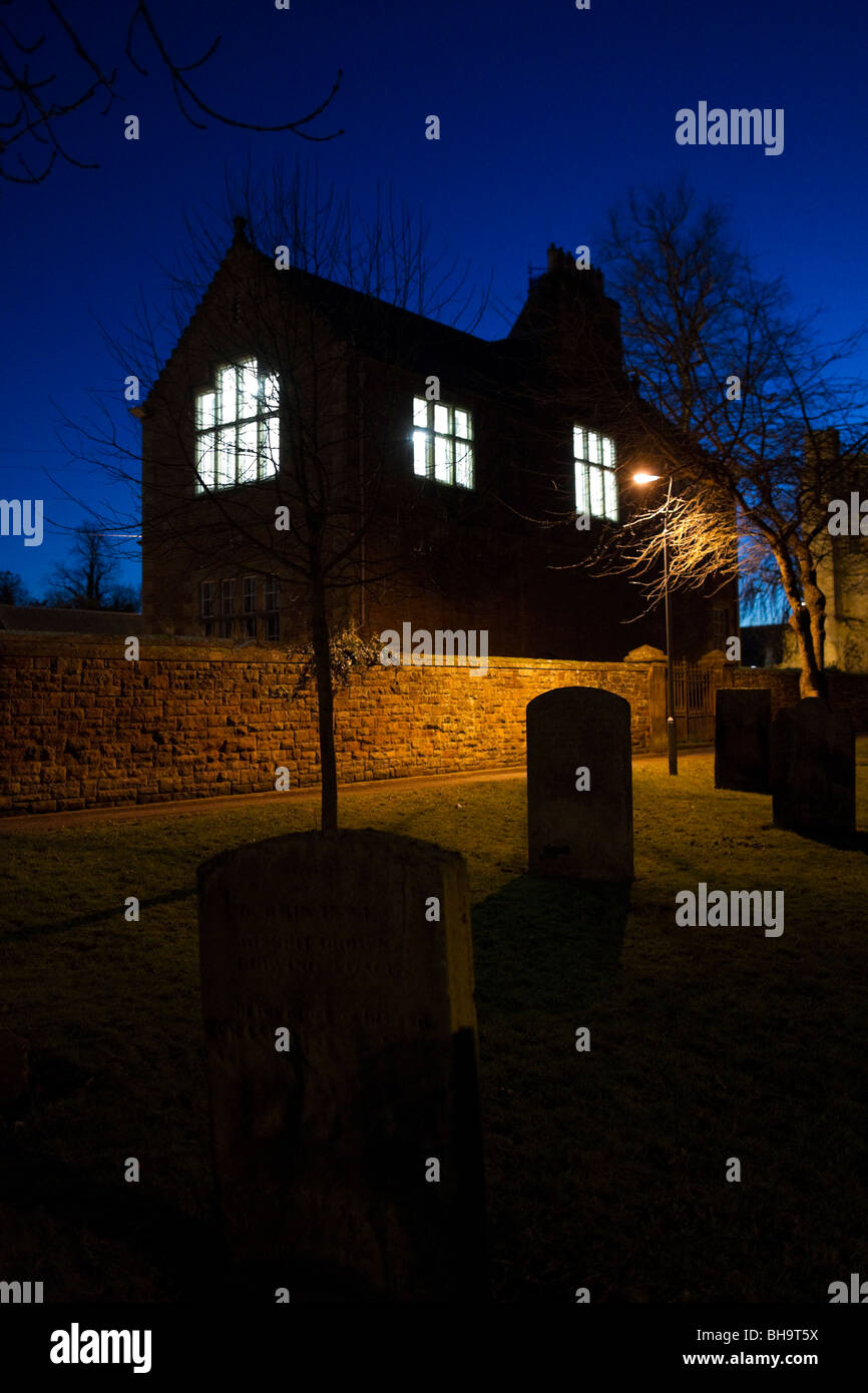 Spooky old schoolhouse with lit up windows at dusk with gravestones in the foreground Stock Photo