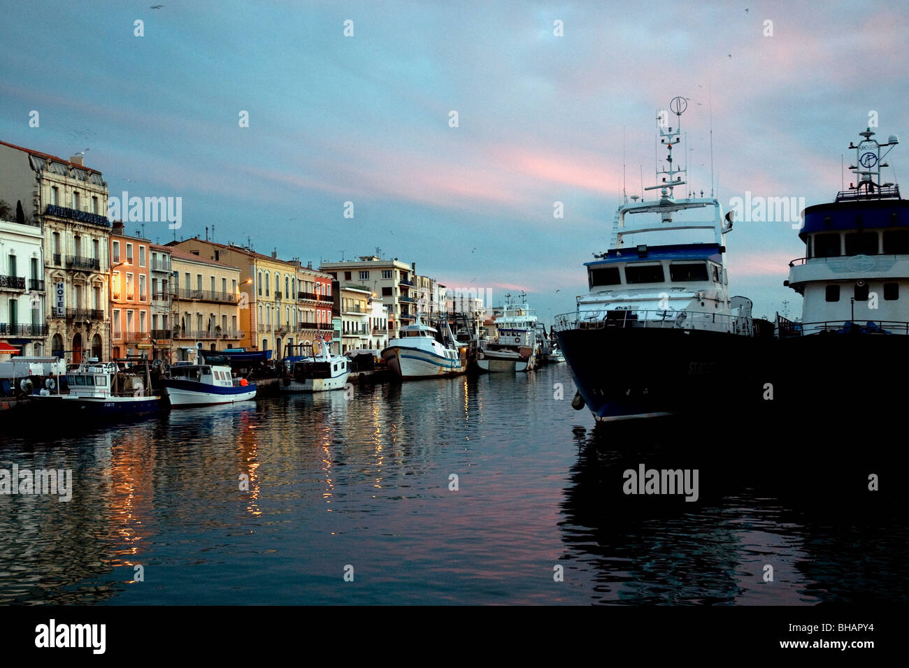 In Sète. France's largest Mediterranean fishing port, buildings glow at sunset beside trawlers moored in the Royal Canal Stock Photo