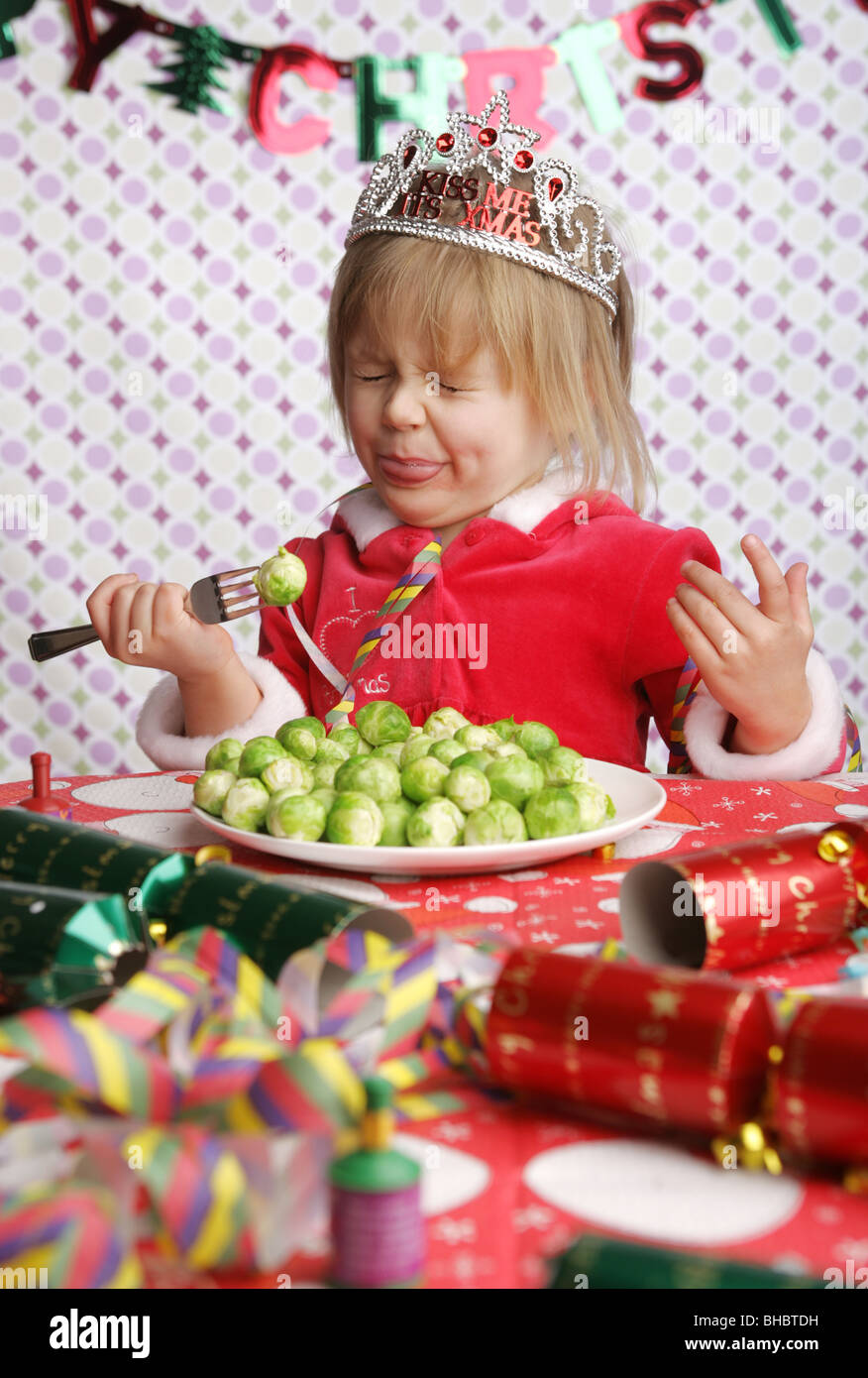 A three year old sitting at a table with Christmas decorations and a plate full of sprouts pulling a face in disgust. Stock Photo