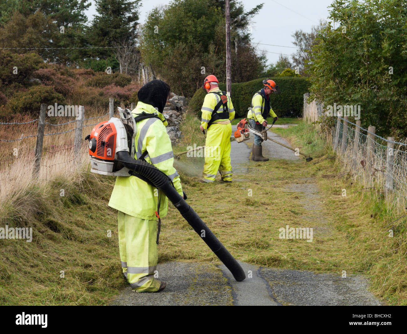 UK, Europe. Workmen wearing hi-visibility suits strimming a grass verge on a country lane Stock Photo