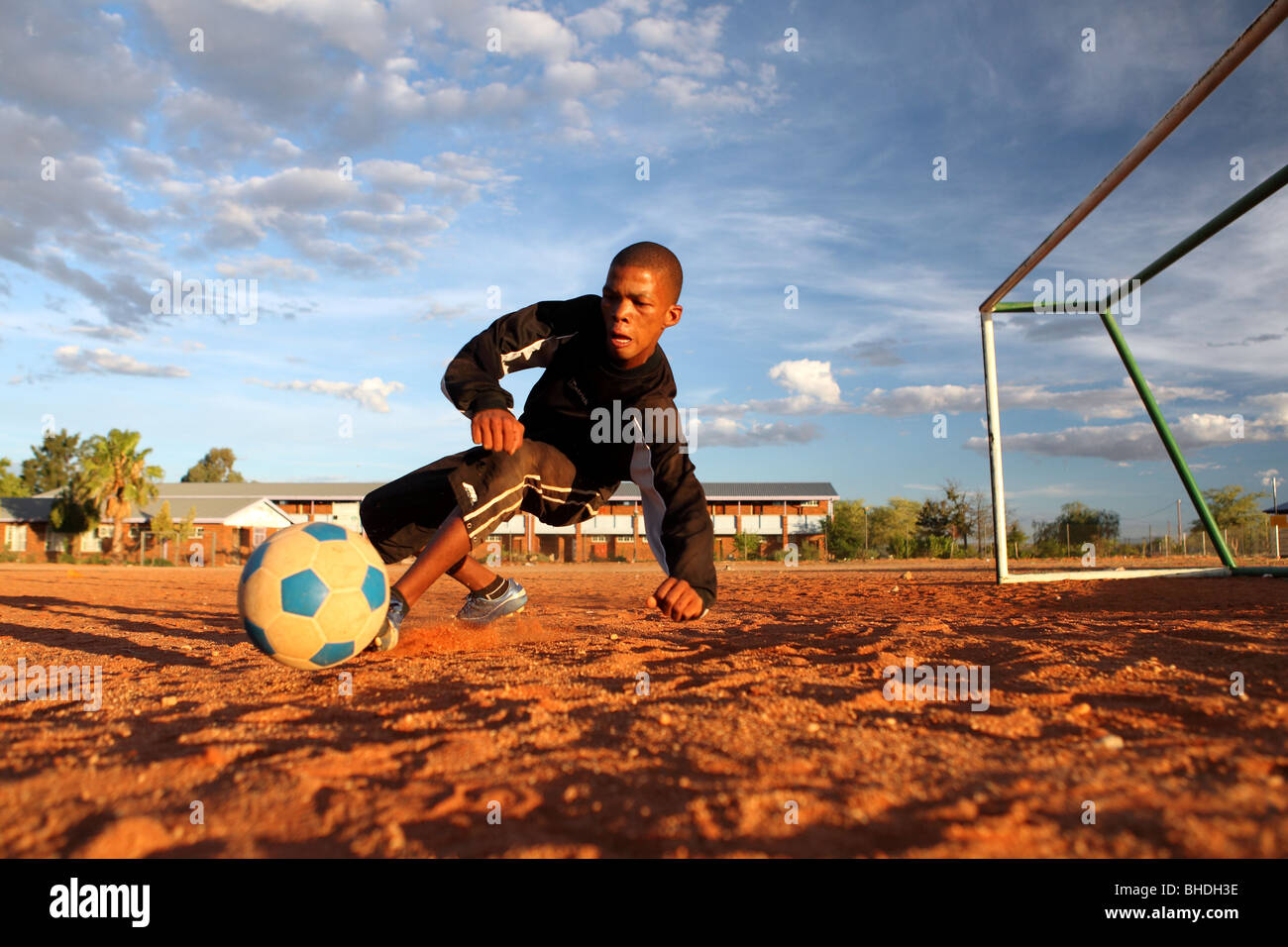 A soccer goalkeeper dives to save the shot at goal on a sandy pitch in
