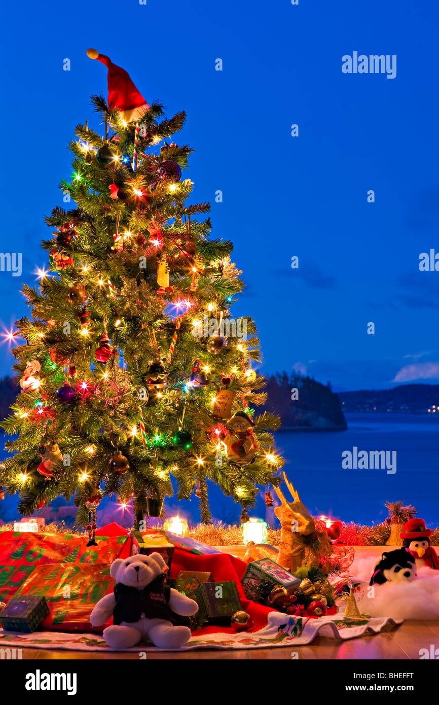 Christmas Tree with lights, decorations, and gifts in a window at dusk, The Artists Point, Hyde Creek, Port McNeill, BC Stock Photo