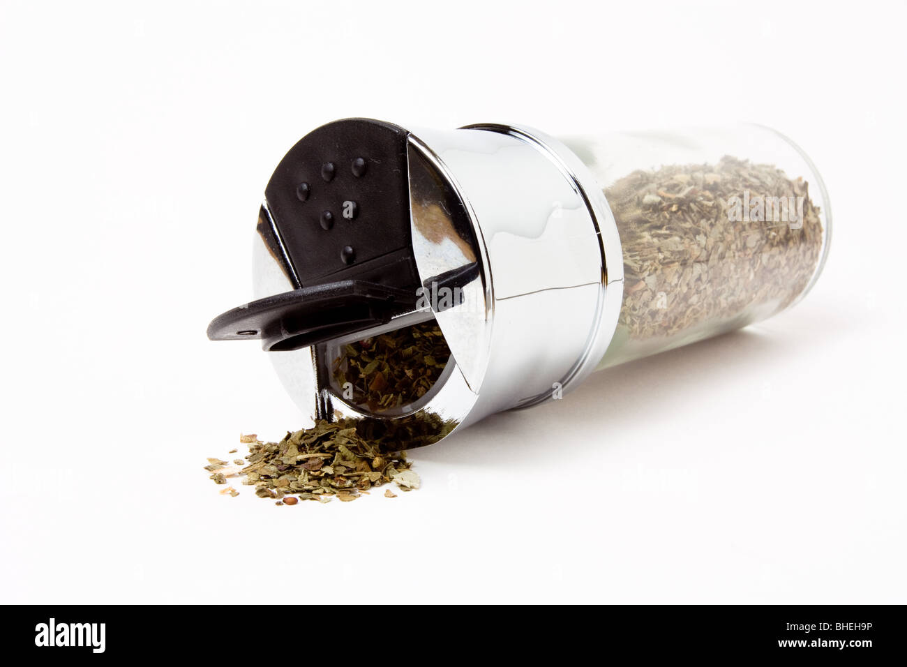 Dried basil spilling from chrome topped dispenser against white background. Stock Photo