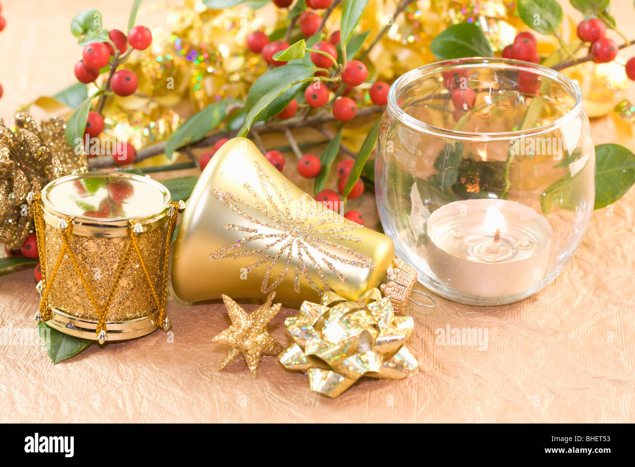 gold bell shaped ornament, drum, star, bow with tea light candle and holly branch on textured gold paper Stock Photo