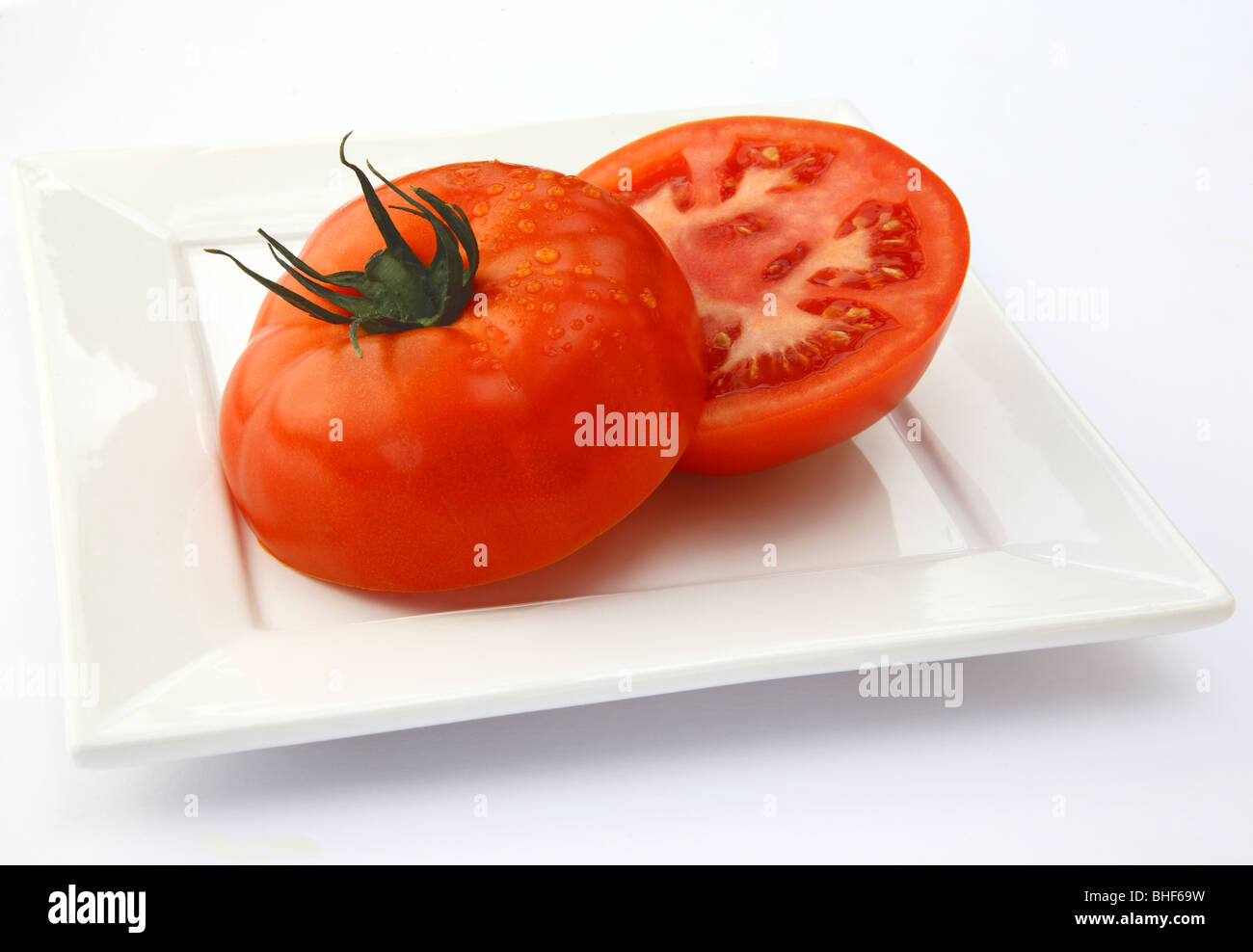 Large, ripe, red, juicy organic beef tomato cut into two halves, on square white plate with white background. Stock Photo