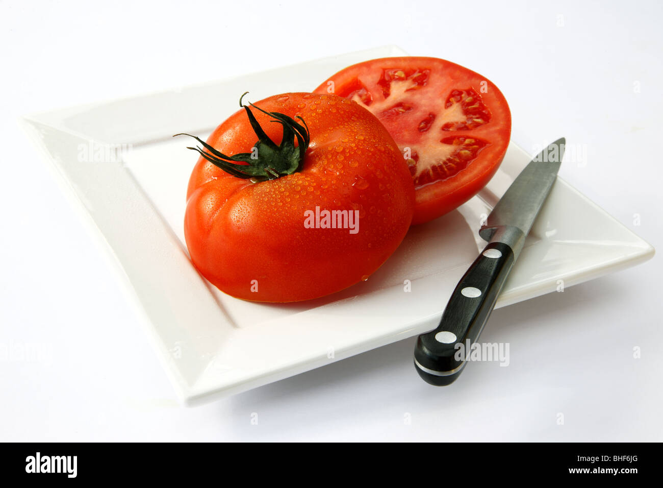 Large, fresh, red, ripe, juicy beef tomato cut into two halves on square white plate with kitchen knife, white background. Stock Photo