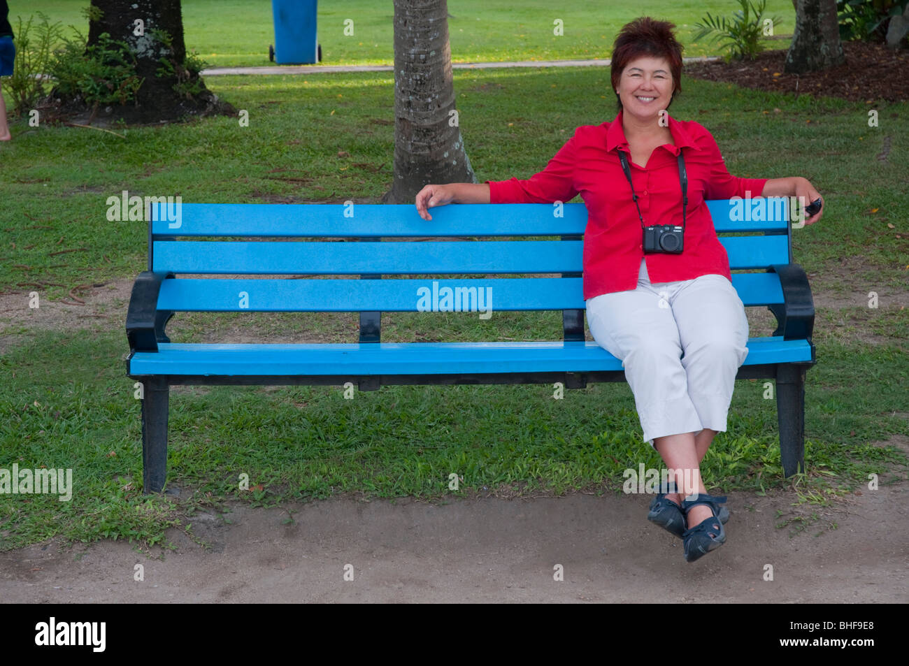 A mature woman in a red shirt on holiday sitting on a blue bench with a Canon G11 camera Stock Photo