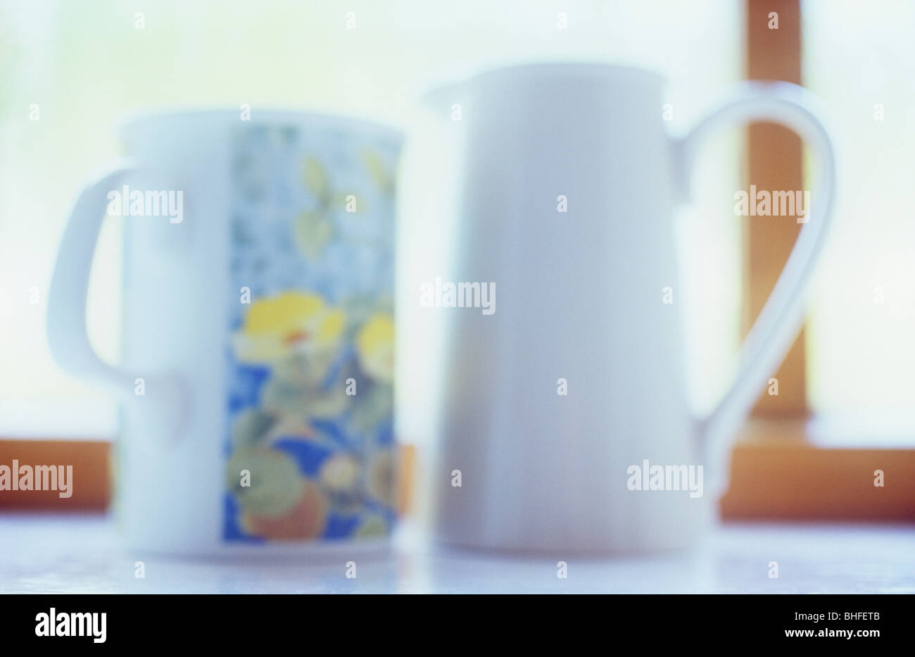 Impressionistic close up at eye level of mug with floral pattern and white milk jug on windowsill with window behind Stock Photo