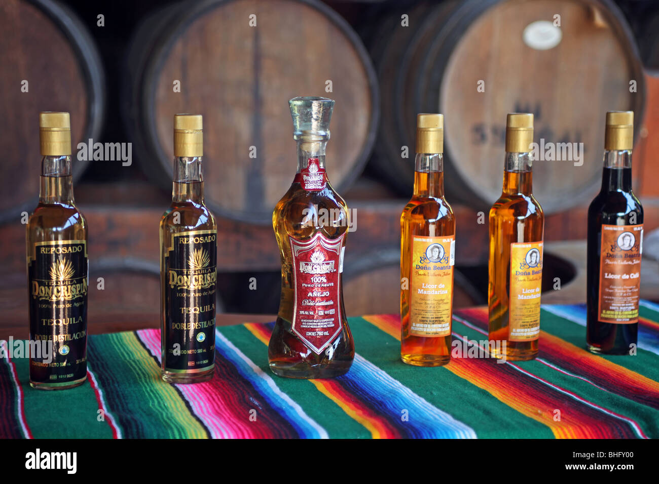 Tequila alcohol in bottles at distillery near Puerto Vallarta, Mexico. Colorful bottles on Mexican blanket in front of barrels. Stock Photo