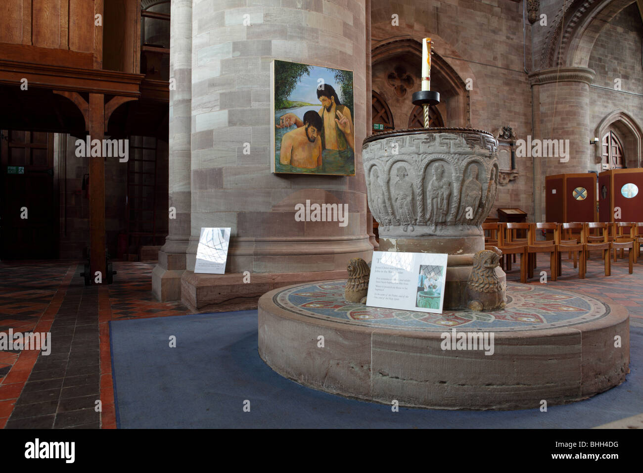 Set upon a circular plinth,the stone carved Font at Hereford Cathedral set against a picture of a Baptism hung on a huge column. Stock Photo