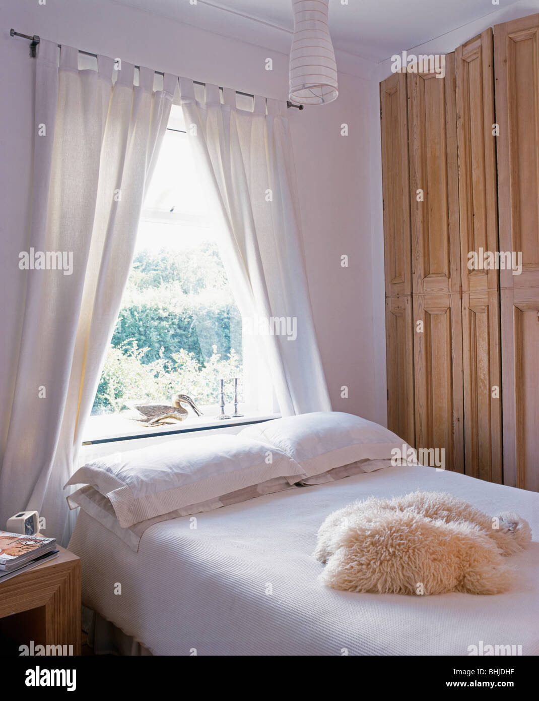 White curtains on window above bed with bedlinen and sheepskin throw in white bedroom with folding wooden screen Stock Photo