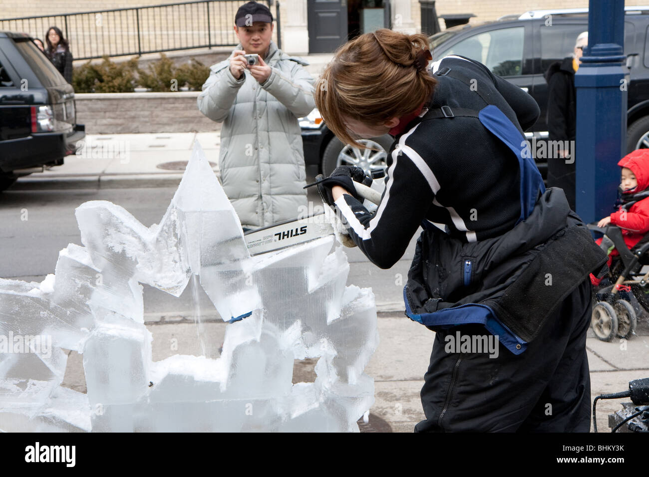 An ice carving artist demonstrating her skill while a bystander looks on Stock Photo