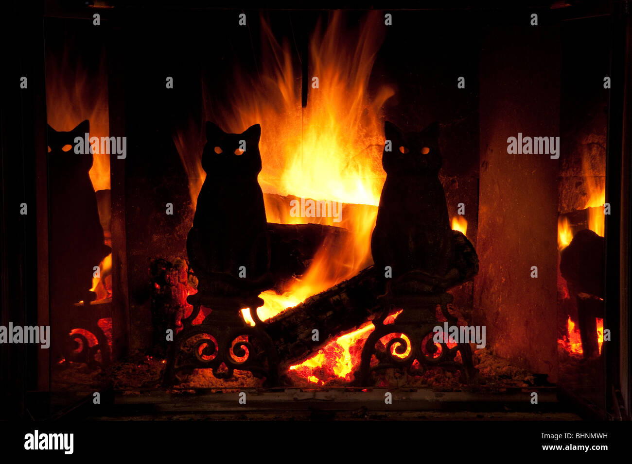 Brilliant orange and yellow flames behind silhouetted black cat figure cast iron andirons on hearth in  fireplace, big fire glows through eyes of cats Stock Photo