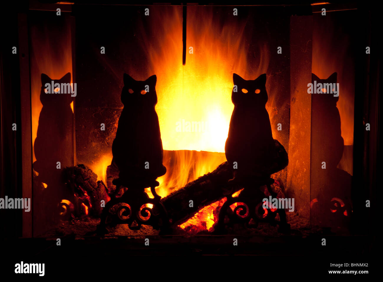 Brilliant yellow and orange flames behind silhouetted black cat wrought  iron andirons in cozy warm fireplace. Flames glow through eyes of cats Stock Photo