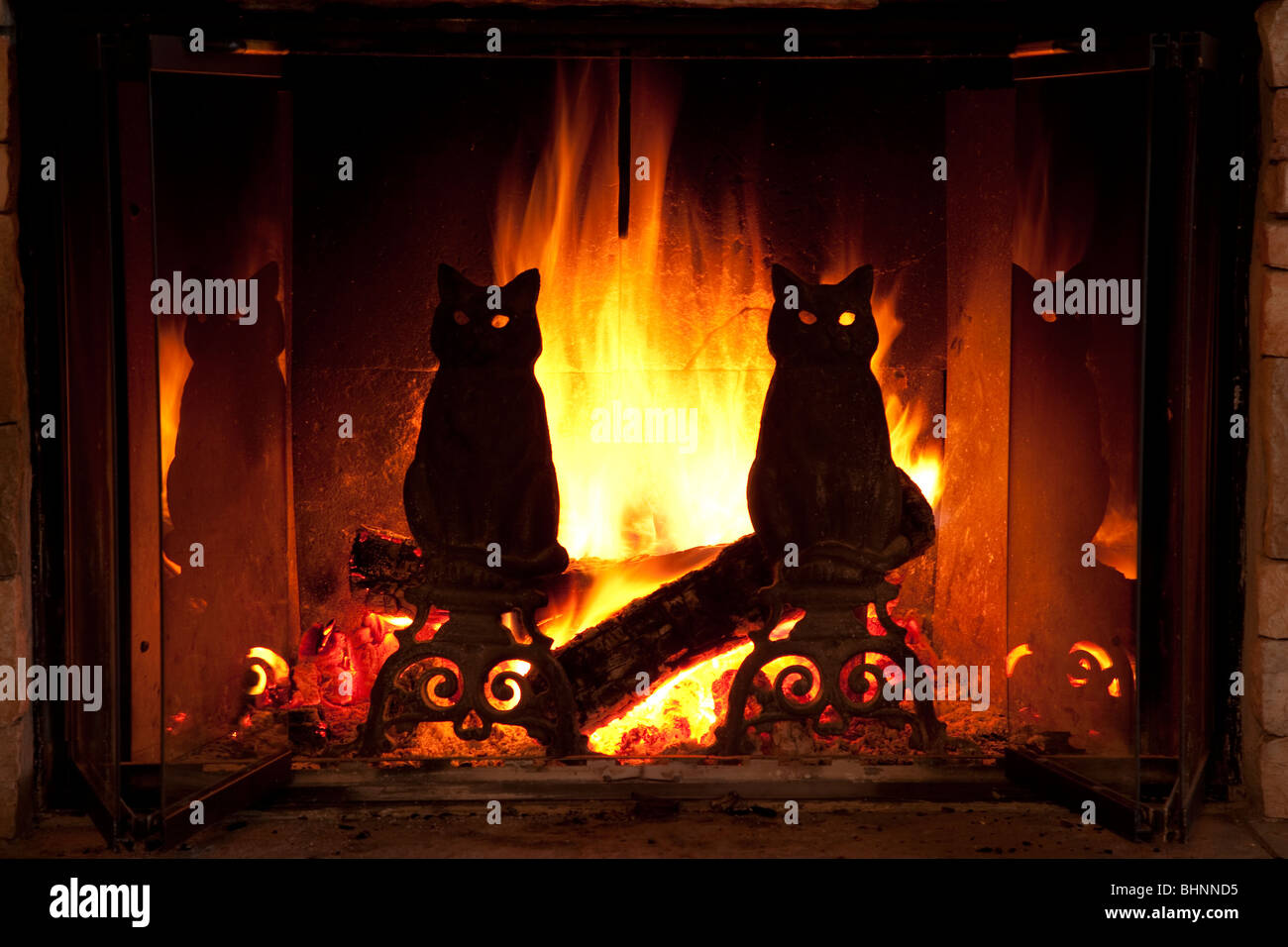 Brilliant yellow and orange flames behind silhouetted black cat andirons in open wood burning fireplace. Flames glow through eyes of cat figures Stock Photo